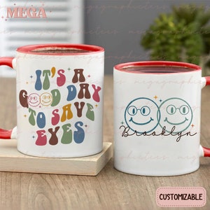 May include: Two white ceramic mugs with red rims. One mug has the text "It's a good day to save your eyes" in colorful retro font with smiling faces. The other mug has two smiling faces with glasses and the name "Brooklyn" written below. Both mugs are on a wooden surface.