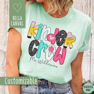 Può includere: T-shirt verde chiaro con la scritta "Kinder Crew" in un design colorato e giocoso. Il design include una matita, un cuore, una mela e il nome "Ms. Williams". La maglietta è etichettata "Bella Canvas" e "Personalizzabile".
