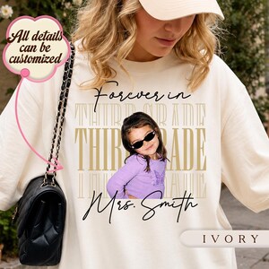 Może przedstawiać: Bluza w kolorze kości słoniowej z nadrukiem graficznym zawierającym tekst Forever in THIRD GRADE oraz Mrs. Smith. Projekt zawiera zdjęcie dziecka w fioletowej bluzce z długim rękawem i czarnych okularach przeciwsłonecznych na tle powtarzającego się tekstu.