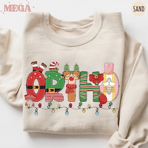 Könnte beinhalten: Ein sandfarbener Pullover mit dem Wort "ORTHO" in einem festlichen Weihnachtsdesign. Die Buchstaben sind mit festlichen Elementen wie Weihnachtsmannbeinen, Weihnachtsbäumen und einem Nussknacker verziert. Eine Lichterkette in bunten Farben verläuft am unteren Rand.