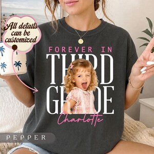 Może przedstawiać: Ciemnoszary t-shirt z białym napisem "FOREVER IN THIRD GRADE" oraz różowym imieniem "Charlotte" pod zdjęciem dziecka z kręconymi włosami, które wystawia język. Różowa dymek z tekstem głosi "All details can be customized."