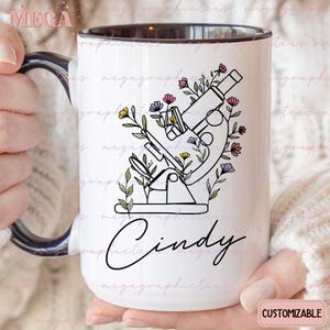 Può includere: Una tazza in ceramica bianca con un manico nero. La tazza presenta un disegno in bianco e nero di un microscopio con dettagli floreali. Il nome "Cindy" è scritto in corsivo sotto l'immagine.
