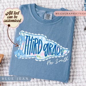 Op de afbeelding: Een blauw spijkerkleurig t-shirt met een afbeelding van een wimpel met een lichtblauw vichy-ruitjes- en bloemenpatroon. De tekst luidt "THIRD grade" in blauw handschrift, met "Mrs. Smith" eronder. Twee lichtblauwe strikken sieren de linkerkant.