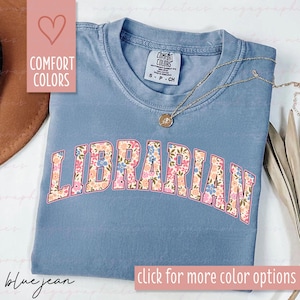 Op de afbeelding: Een blauw spijkerbroekkleurig t-shirt met het woord "LIBRARIAN" in een bloemenontwerp over de voorkant. De shirt heeft een ronde hals en korte mouwen. Een gouden ketting met een hanger is over het shirt gedrapeerd. De woorden "COMFORT COLORS" staan op een roze hart.