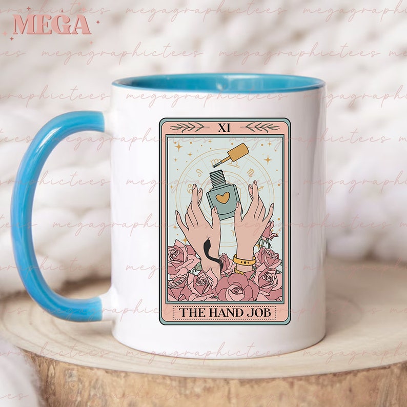 May include: A white ceramic mug with a light blue rim features a tarot card design. The card is titled "The Hand Job" and depicts two hands holding a bottle of nail polish with a heart-shaped design. The card is surrounded by pink roses.