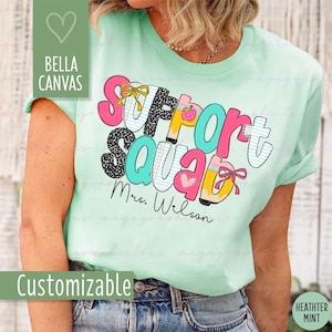 May include: A mint green t-shirt with the words "Support Squad" in colorful, playful lettering. The design includes a strawberry, pencil, and heart. The text "Mrs. Wilson" is written below the main text. The shirt is from Bella Canvas and is customizable.