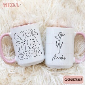 May include: Two white ceramic mugs with pink rims. One mug has the text "Cool Tia Club" in black outlined letters. The other mug has a black line drawing of a flower and the text "Jennifer".
