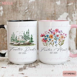 May include: Two white ceramic mugs with black handles. The mug on the left has a mountain scene with the text "Foster Dad - EST. 2025". The mug on the right has a floral design with the text "Foster Mom - EST. 2025".