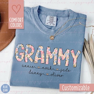 May include: A blue t-shirt with the word "GRAMMY" in large, floral letters. Below the word are the names "annie", "noah", "jade", "danny", and "oliver" with hearts between each name. The shirt is customizable.