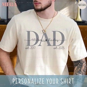 Op de afbeelding: Een wit T-shirt met het woord "DAD" in grijze letters. De tekst "Wilson Harley" staat onder het woord "DAD" met een klein hartje tussen de namen. De tekst "est. 2017" staat onder de linkerkant van het woord "DAD" en de tekst "est. 2025" staat onder de rechterkant van het woord "DAD".