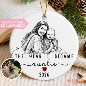 May include: A round white ceramic ornament featuring a black line-art illustration of an adult holding a baby. Text reads: THE YEAR I BECAME auntie 2026. A small circular inset shows a color photograph of the same subjects. A gold string is attached.
