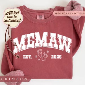 Può includere: Una felpa girocollo color cremisi con la scritta bianca stampata MEMAW EST. 2026, accompagnata da due impronte di piedi di neonato con cuori e piccoli fiocchi di neve. Un fumetto nell'angolo riporta All text can be customized.