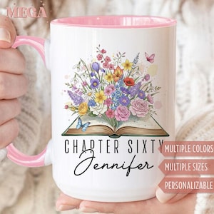 May include: White ceramic mug with a pink interior and handle. The mug features a floral design with a book and butterflies. Text on the mug reads "Chapter Sixty Jennifer". The mug is held by a person.