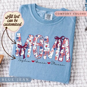 Può includere: Una maglietta Comfort Colors "Blue Jean" azzurra con la scritta "MOM" in un motivo floreale e a bandiera americana. I nomi Sophia, Grace e Steven sono sotto la parola "MOM". Il testo "All text can be customized" è in una nuvoletta.