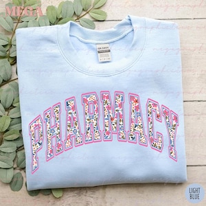 Pu&ograve; includere: Felpa blu chiaro con la parola "FARMACIA" in lettere bianche bordate di rosa. Le lettere sono riempite con un motivo floreale rosa, blu e verde.