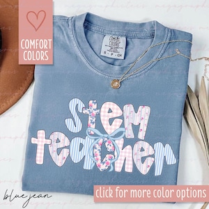 May include: A blue jean Comfort Colors t-shirt with the words "STEM Teacher" in a playful font. The letters are decorated with floral, gingham, and striped patterns. A gold necklace with a pendant rests on the shirt. The image also includes text that says "click for more color options."