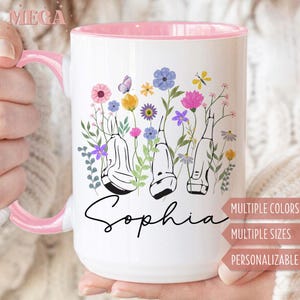 May include: White ceramic mug with a pink interior and handle. The mug features a floral design with colorful flowers, butterflies, and the name "Sophia" in black script. Text on the side indicates multiple colors, sizes, and personalization options.