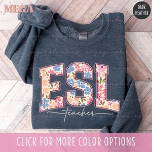 May include: Dark heather gray sweatshirt with the word "ESL" in a floral design, with the word "teacher" underneath. The sweatshirt has a crew neck and long sleeves. The text "CLICK FOR MORE COLOR OPTIONS" is at the bottom.