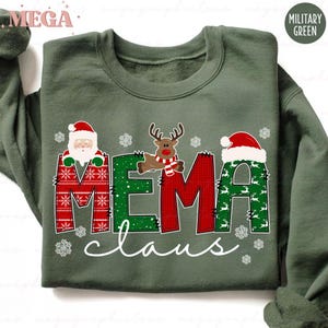 Könnte beinhalten: Militärgrünes Sweatshirt mit dem Text "MEMA Claus" in Rot und Grün. Die Buchstaben sind mit dem Weihnachtsmann, einem Rentier und Schneeflocken verziert. Ein festliches Kleidungsstück für die Feiertage.