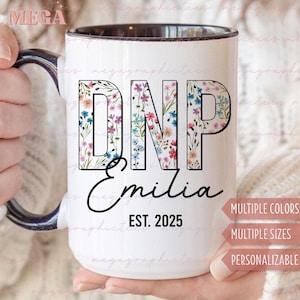 Può includere: Una tazza in ceramica bianca con bordo e manico neri. La tazza presenta un motivo floreale con le lettere "DNP" e il nome "Emilia" con il testo "EST. 2025" sotto.