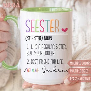 May include: White ceramic mug with a green handle and interior, featuring the word "SEESTER" in colorful letters. The mug includes definitions and the name "Jadie". The mug is held by a person.