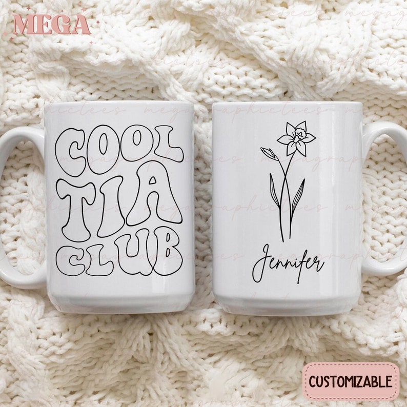 May include: Two white ceramic mugs with black designs. One mug has the text "Cool Tia Club" in a retro font. The other mug has a simple line drawing of a daffodil and the name "Jennifer".