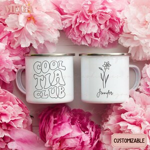 May include: Two white enamel mugs with black designs. One mug says "Cool Tia Club" in a retro font. The other mug has a simple line drawing of a daffodil and the name "Jennifer" written below it.