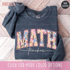 May include: Dark heather gray sweatshirt with the word "MATH" in a floral design, and the word "Teacher" in a script font. The sweatshirt has a crew neck and long sleeves. The text "CLICK FOR MORE COLOR OPTIONS" is at the bottom.