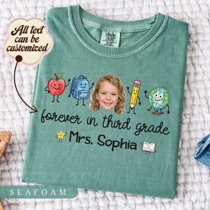 Może przedstawiać: Morski zielony t-shirt z nadrukiem uśmiechniętego jabłka, plecaka, ołówka i globusa z twarzą dziecka pośrodku. Tekst brzmi "forever in third grade" oraz "Mrs. Sophia" wraz z małą gwiazdką i ikoną otwartej książki na koszulce z krótkim rękawem.