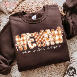 Könnte beinhalten: Dunkelbraunes Sweatshirt mit dem Wort "MEMAW" in einem herbstlichen Design. Die Buchstaben sind mit Karomustern und Kürbisgrafiken gefüllt. Unter dem Wort stehen die Namen "Oliver", "Mia" und "Sophie".