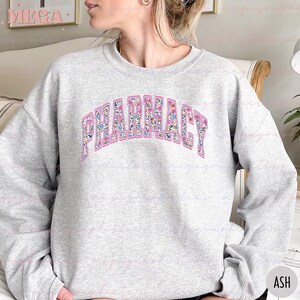 Pu&ograve; includere: Una felpa grigia con la parola "FARMACIA" stampata in lettere rosa con un motivo floreale. Le lettere sono in un font in stile universitario.