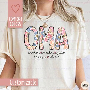 Puede incluir: Camiseta color crema con la palabra "OMA" en letras grandes y llenas de flores. Debajo est&aacute;n los nombres "annie", "noah", "jade", "danny" y "oliver". La camiseta tambi&eacute;n tiene las palabras "Comfort Colors" y "Customizable" en el lado izquierdo.
