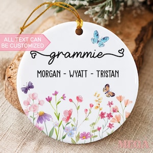 May include: A round white ceramic ornament featuring a gold hanging string, decorated with colorful watercolor wildflowers and butterflies. The text reads "grammie" in script and "MORGAN - WYATT - TRISTAN" in block letters, with a pink label stating "ALL TEXT CAN BE CUSTOMIZED".