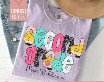 Puede incluir: Una camiseta de color lila claro con las palabras "Second Grade" en una fuente colorida y juguetona. El diseño incluye lápices, un clip y una manzana. El texto "Mrs. William" está debajo. La camiseta es parte de la colección "Comfort Colors".