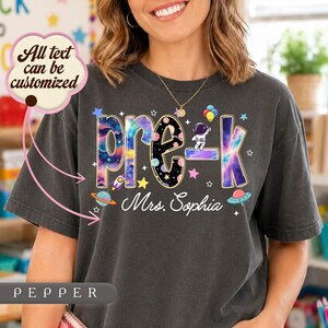 May include: A charcoal gray short-sleeve t-shirt featuring the text pre-k in a colorful space-themed font with planets, stars, and an astronaut. Below the text, the name Mrs. Sophia is printed in white script. A text bubble reads All text can be customized.