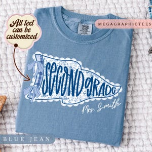 May include: A blue jean coloured t-shirt featuring a white pennant graphic with a blue floral pattern and lace trim. The text reads Second Grade and Mrs. Smith. A blue plaid ribbon with two bows is attached to the left side of the pennant graphic.