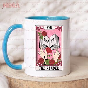 May include: White ceramic mug with a light blue rim and handle. The mug features a pink and white tarot card design with the number XVIII and the words "SMUT" and "THE READER".