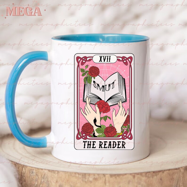 May include: White ceramic mug with a light blue rim and handle. The mug features a pink and white tarot card design with the number XVIII and the words "SMUT" and "THE READER".