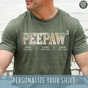 May include: Heather military green t-shirt with the word "PEEPAW" in a map-like font. Below the word are the names Jadie, Cadie, and Sadie with their established years. The shirt also has the text "PERSONALIZE YOUR SHIRT".