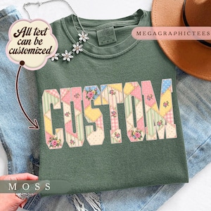 Pode incluir: Uma t-shirt verde musgo com a palavra CUSTOM numa fonte estilo patchwork com padrões florais, de bolinhas e listrados em rosa, amarelo, azul e verde. A t-shirt inclui uma etiqueta Comfort Colors e o texto All text can be customized.