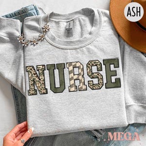 May include: An ash grey crewneck sweatshirt featuring the word NURSE in block letters. The letters have a mixed pattern design including camouflage, olive green, and a checkered print. A silver-toned floral necklace rests on the fabric.