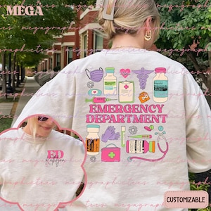 May include: A light gray sweatshirt with a pink and blue graphic design of medical supplies and the text "EMERGENCY DEPARTMENT". The design includes a stethoscope, a syringe, a thermometer, a pill bottle, a first aid kit, and a pair of gloves. The sweatshirt is customizable.