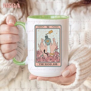 May include: A white ceramic mug with a green rim features a tarot card design. The card depicts a pair of hands holding a bottle of nail polish with a heart on the label. The card is titled "The Hand Job" and has the Roman numeral "XI" in the top corner.
