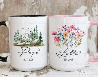 May include: Two white ceramic mugs. One features a mountain and forest scene with the word "Popo" and "-EST. 2025-". The other mug has a floral design with the word "Lolli" and "-EST. 2025-". Both mugs have a heart symbol.