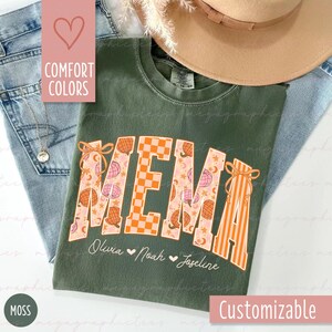 Könnte beinhalten: Ein moosgrünes T-Shirt mit dem Wort "MEMA" in einer dekorativen Schriftart, mit einer Mischung aus Mustern, darunter Kürbisse und Schachbrettmuster. Die Namen Olivia, Noah und Joseline sind darunter aufgedruckt. Das T-Shirt ist anpassbar.
