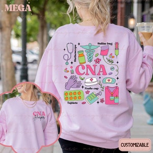 Op de afbeelding: Roze sweatshirt met een grafisch ontwerp van medische instrumenten en de tekst "CNA". Het ontwerp bevat een stethoscoop, een spuit, een zoutoplossingzak, een pilfles, een verband, een verpleegstersmuts en een paar handschoenen. Het sweatshirt is aanpasbaar.