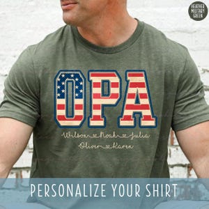 May include: Heather military green t-shirt with the word "OPA" in a patriotic design, featuring the American flag. Below the word "OPA" are names: Wilson, Noah, Julia, Oliver, and Karen. The text "PERSONALIZE YOUR SHIRT" is at the bottom.