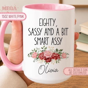 May include: A 15oz white ceramic mug with a pink handle and interior. The front features black text reading EIGHTY, SASSY AND A BIT SMART ASSY, a floral illustration in shades of red and pink with green leaves, and the name Olivia in script.