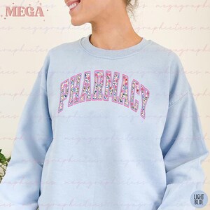 Pu&ograve; includere: Felpa blu chiaro con la parola "FARMACIA" in lettere rosa con un motivo floreale. Le lettere sono contornate di blu e rosa.