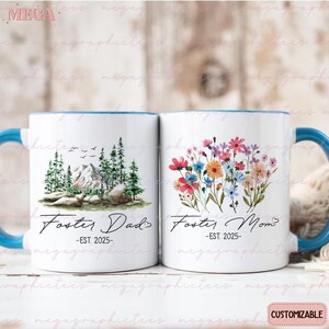 K&ouml;nnte beinhalten: Zwei wei&szlig;e Keramikbecher mit blauen R&auml;ndern. Der Becher links zeigt eine Berglandschaft und den Text "Foster Dad -EST. 2025-". Der Becher rechts hat einen Strau&szlig; bunter Blumen und den Text "Foster Mom -EST. 2025-".